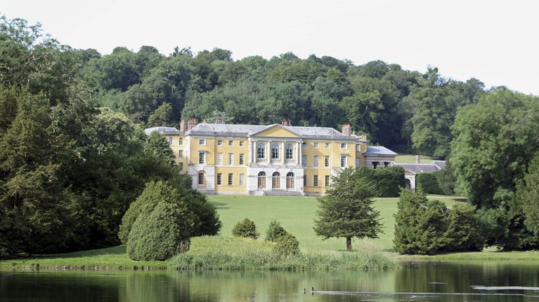 The three storey, yellow stuccoed, Palladian mansion seen from across the lake framed by woods at West Wycombe Park, Village and Hill, Buckinghamshire